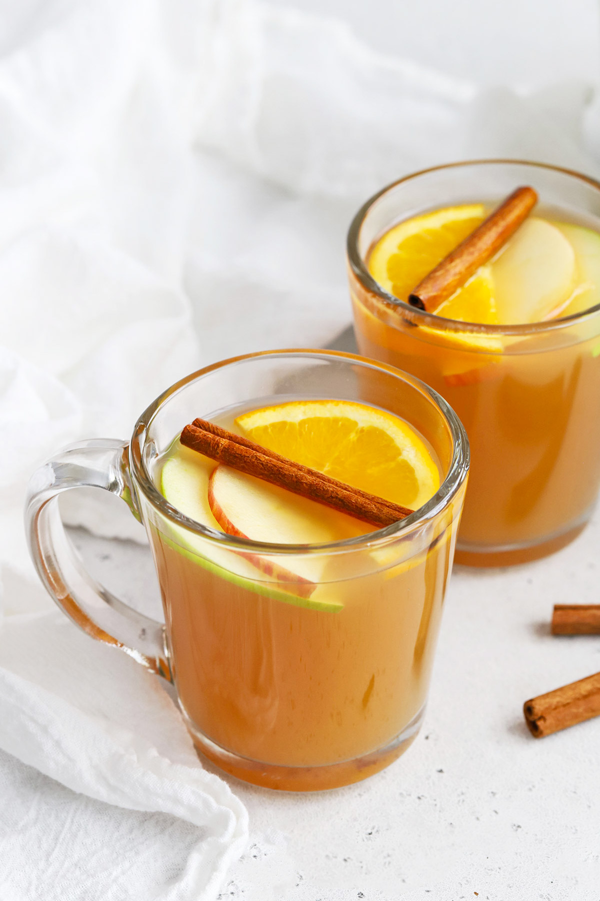 Front view of glass mugs of hot spiced cider garnished with apple slices, orange slices, and cinnamon sticks
