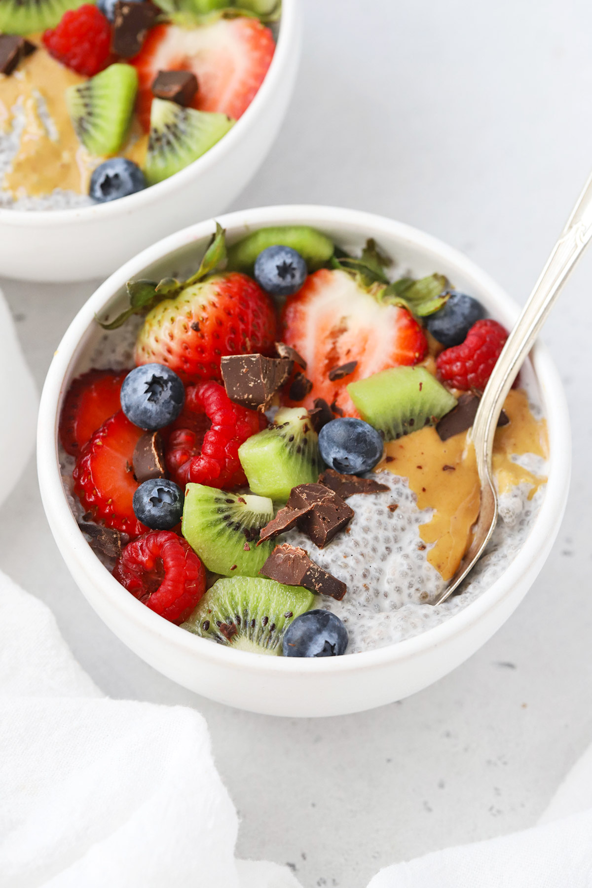 Front view of a white bowl of vanilla chia pudding topped with kiwi, fresh berries, peanut butter, and chocolate chunks