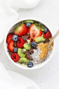 Overhead view of a white bowl of vanilla chia pudding topped with kiwi, fresh berries, peanut butter, and chocolate chunks