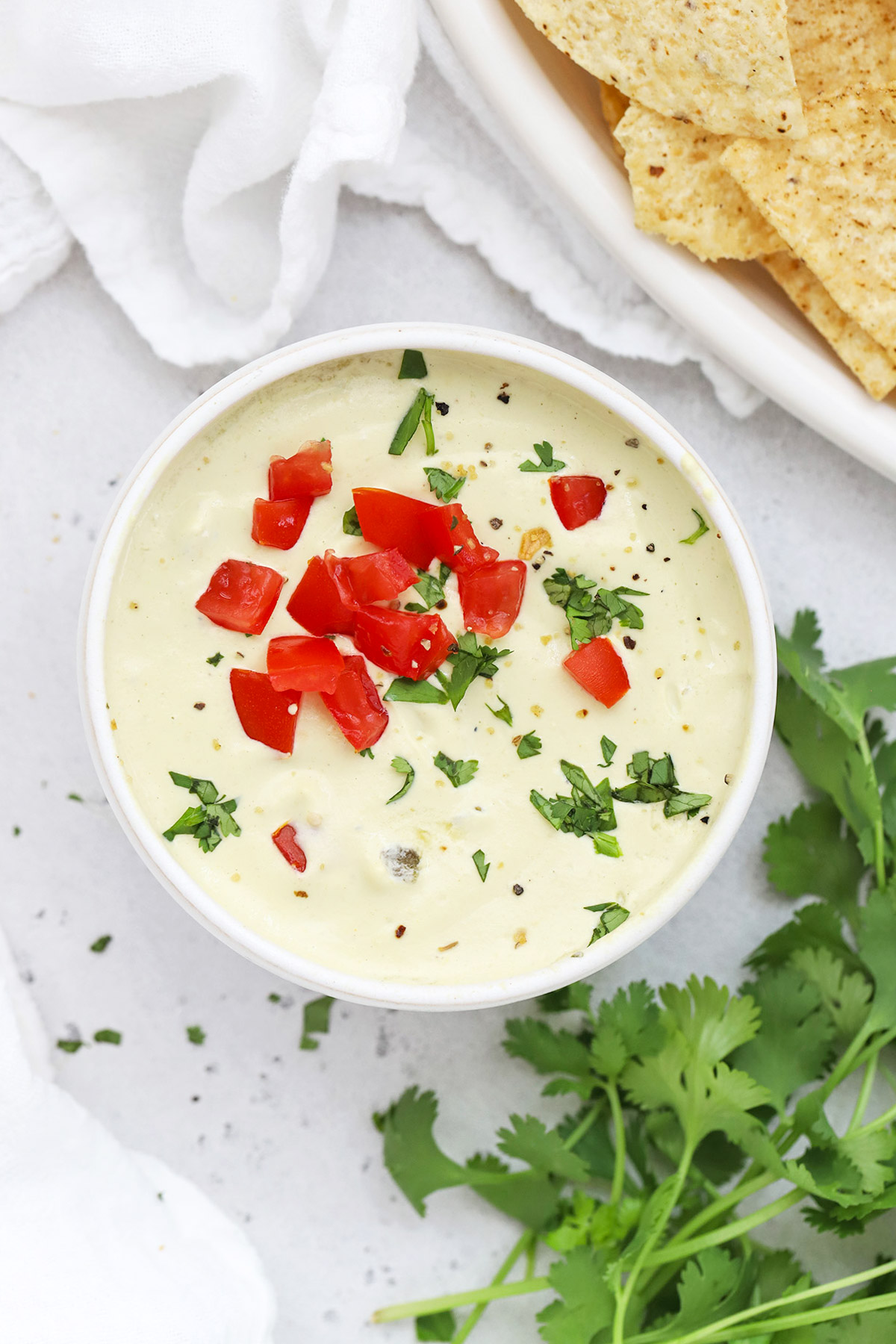 Overhead view of a white bowl of white cashew queso dip topped with chopped tomato and minced cilantro
