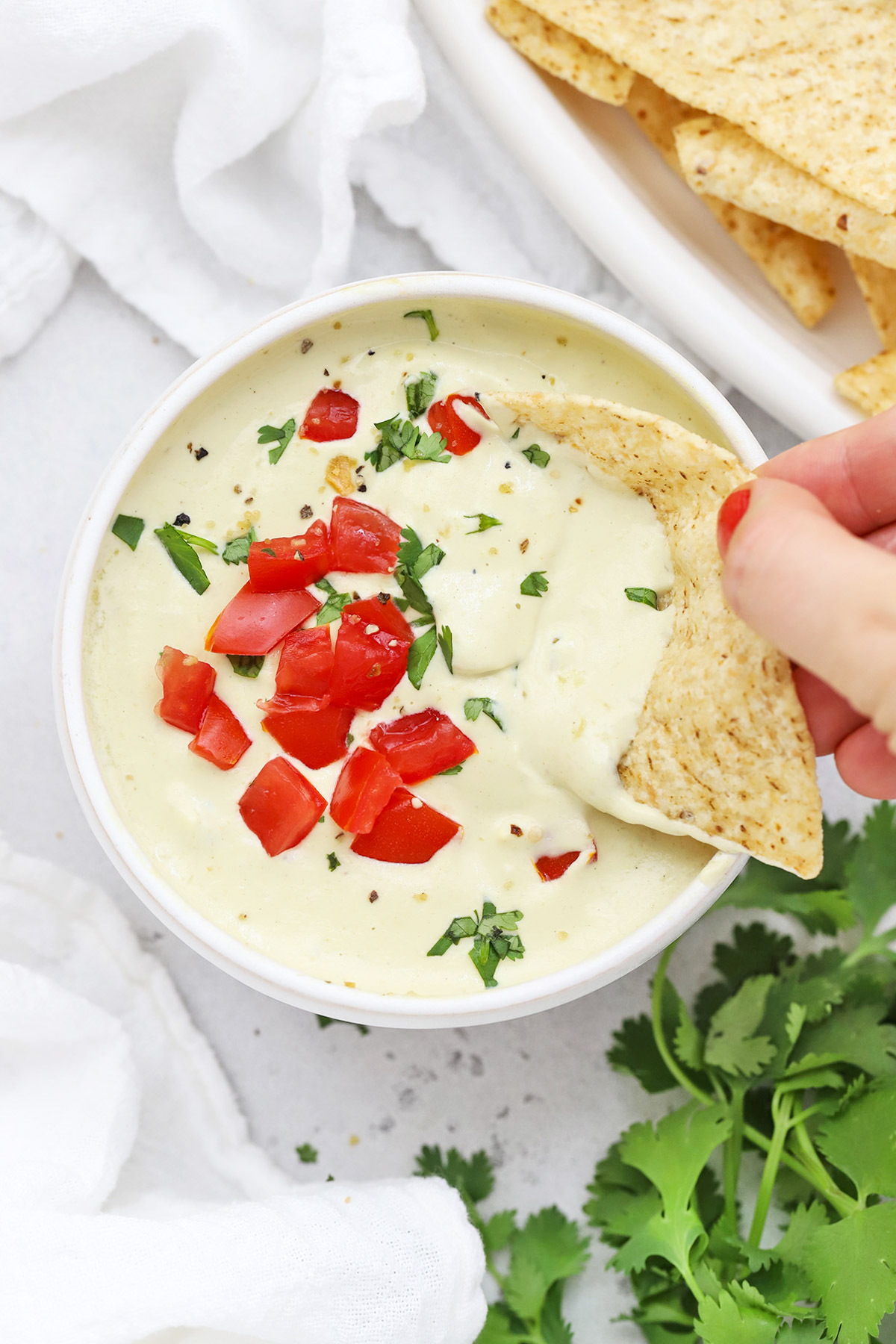 A tortilla chip dipped into a bowl of white cashew queso dip topped with chopped tomato and minced cilantro