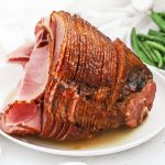 Front view of honey mustard glazed spiral ham on a white platter with a bowl of green beans in the background