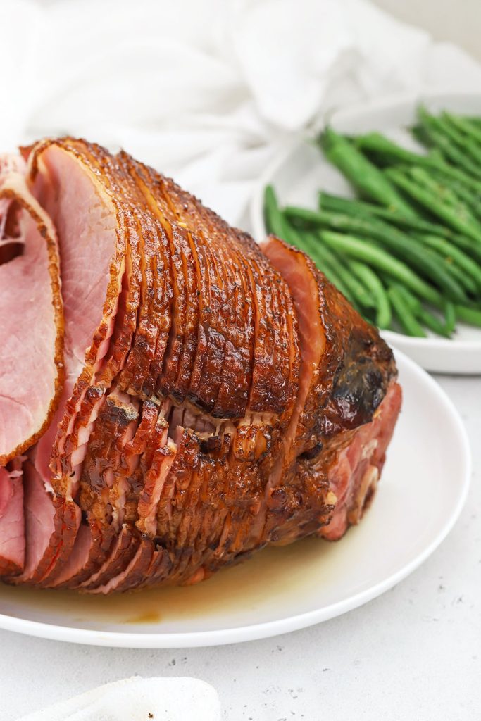 Front view of honey mustard glazed spiral ham on a white platter with a bowl of green beans in the background