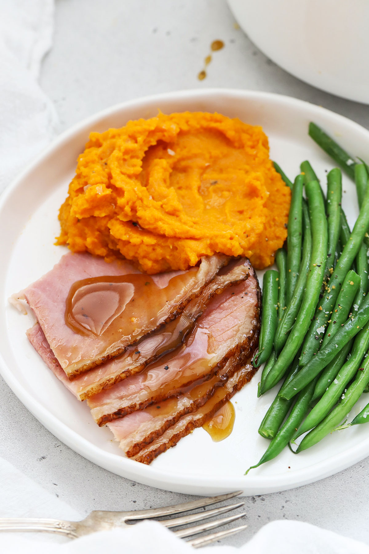 Slices of glazed ham on a white plate with mashed sweet potatoes and steamed green beans