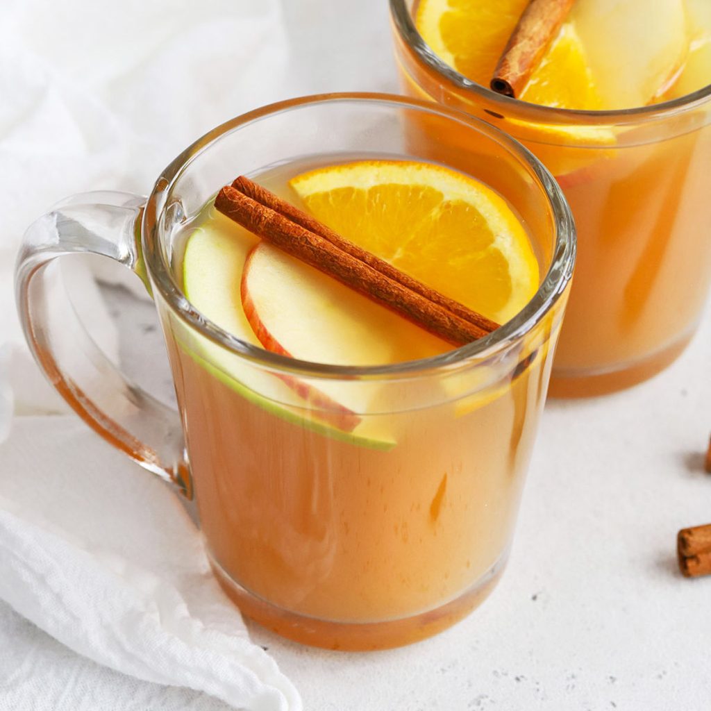 two glass mugs of hot spiced cider with apple slices, orange slices, and cinnamon sticks