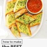 Overhead view of slices of garlic bread on a white platter with a bowl of marinara sauce