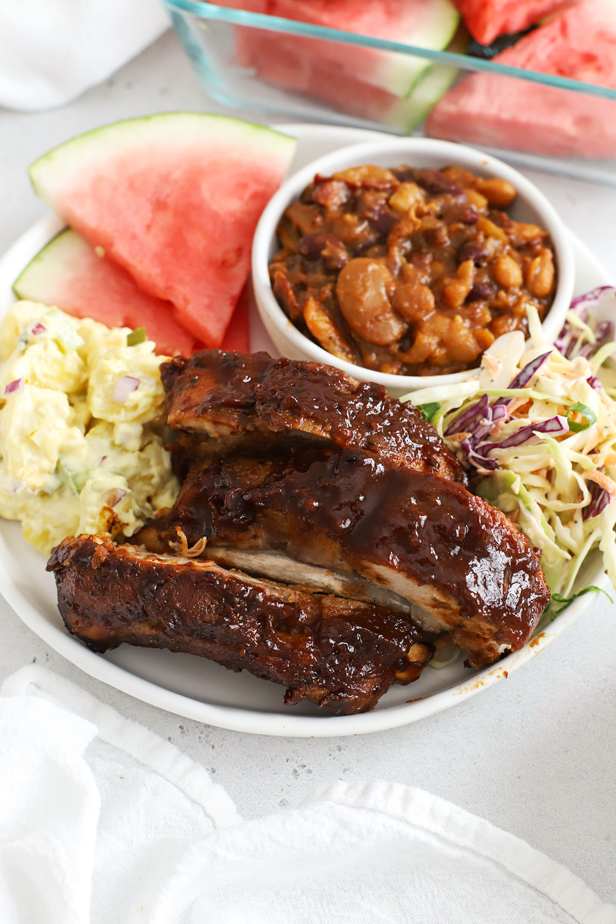 crock pot ribs on a plate with potato salad, coleslaw, watermelon, and baked beans