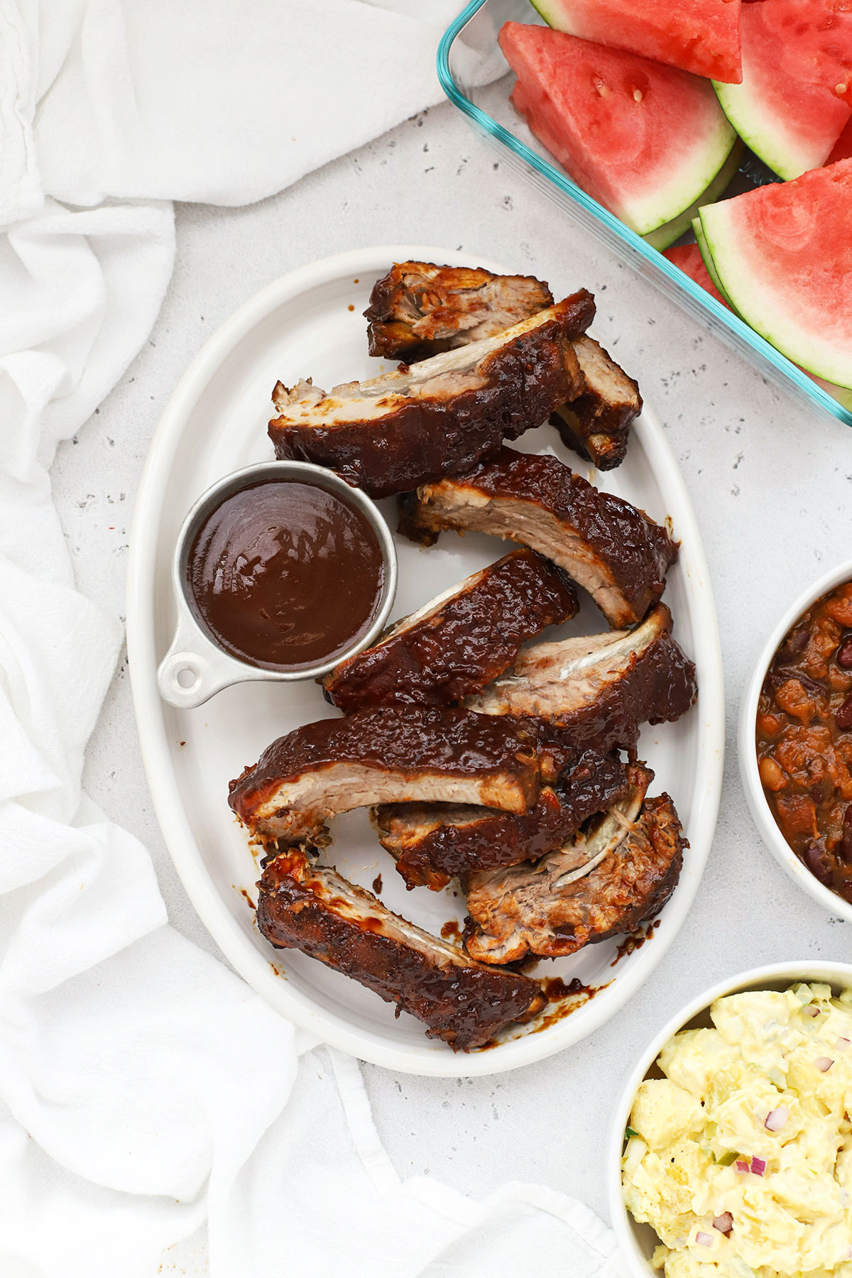 slow cooker ribs on a platter surrounded by bowls of side dishes--watermelon, baked beans, and potato salad