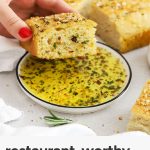 a slice of gluten-free focaccia being dipped into dipping oil with herbs