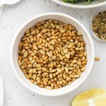 toasted pine nuts in a white bowl next to arugula salad ingredients