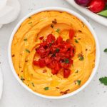 Homemade roasted red pepper hummus in a white bowl with a plate of colorful veggies next to it