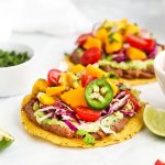 two black bean tostadas topped with guacamole, cilantro lime slaw, fresh mango salsa, and garnishes