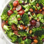 white serving bowl of vegan broccoli salad with red grapes, dried cranberries, toasted almonds, red onions, and poppy seed dressing