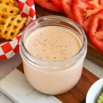 A jar of burger sauce surrounded by burger toppings and a basket of waffle fries