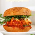 A chicken parmesan burger topped with arugula salad on a white plate